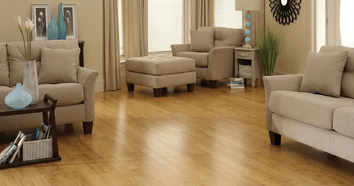 A living room with professionally installed bamboo flooring, beige sofas, a matching ottoman, a side table with a blue lamp, vases, framed pictures, and large windows with blinds and curtains in Atlanta, GA